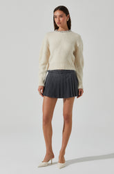 A young woman is wearing a fluffy, cream-colored, long-sleeve sweater with a decorative collar. She pairs it with a high-waisted, dark gray pleated mini skirt that has a waistband with belt loops and a button closure. She is also wearing light-colored high heel shoes.
